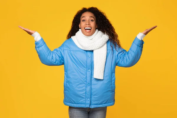 Happy surprised black lady wearing winter jacket and scarf, with mouth open and hands spread, looks at the camera on yellow background, astonished by great offer