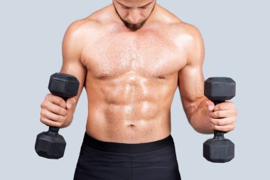 Ciddi terli, güçlü, kaslı, Avrupalı, sakallı, çıplak gövdeli genç sporcu, dambıllarla egzersiz yapıyor, kas çalıştırıyor, gri stüdyo arka planında izole edilmiş, kırpılmış.