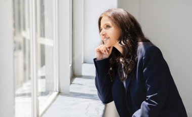 Resmi kıyafet giymiş çekici, olgun bir kadının camdan bakıp gülümsemesinin yan görüntüsü. İş kadını güneşli günün tadını çıkarıyor, ofiste çalışıyor, fotokopi çekiyor.