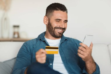 Akıllı telefon ve kredi kartı kullanan mutlu orta yaşlı bir adam. Kanepede oturan müşteri ekommerce başvurusu, bankacılık ve perakende satış yapıyor.