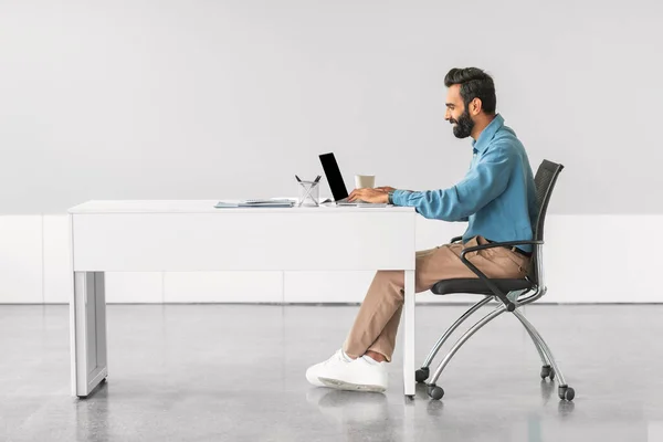 Engaged indian businessman using laptop at minimalist white desk, with coffee and notebook, symbolizing focused work in modern office setting, side view, free space
