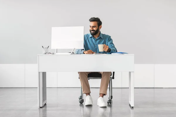 Content indian male professional with beard enjoying his coffee while working on computer at clean and minimalistic white office desk, full length
