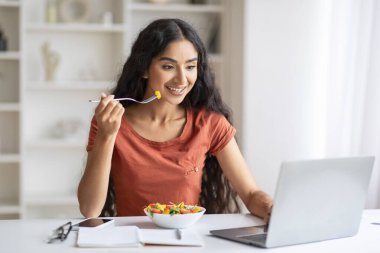 Serbest çalışan genç Hintli bir kadın öğle yemeğini evde yer, ofis masasında oturur, sağlıklı salata yer, dizüstü bilgisayar yer, e-postaları kontrol eder, fotokopi çekerdi. Bağımsızların yaşam tarzı
