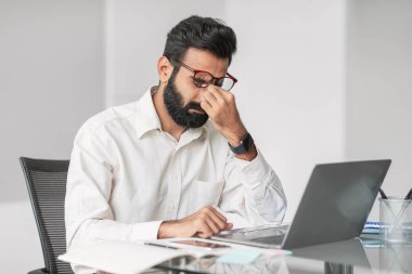 Yorgun Hintli işadamı burnuna masaj yapıyor. Ofis masasında çalışırken stres belirtileri gösteriyor. Uzun çalışma saatlerini ya da iş basıncını yansıtıyor.