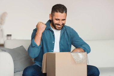 Orta yaşlı bir adam sevinçle oturma odasında teslimat kutusunu açıyor, başarılı online alışverişi kutluyor, gülümsüyor ve siparişiyle karton paketin içine bakıyor.
