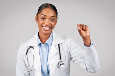 Bin yıllık güzel siyahi kadın doktor ilaç kapsülünü gri arka planda, stüdyoda izole edip kapat. Tıbbi yardım, sağlık tavsiyesi, ilaç, ağrı kesici, vitamin, antidepresan.