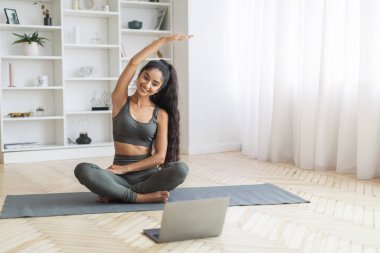 Evde pilates: esmer doğulu kadın içeride egzersiz yapıyor. Sağlıklı yaşam tarzı: Genç Hintli bir kadın yoga minderinde spor yapıyor, dizüstü bilgisayarında iyi aydınlatılmış bir yaşam alanında bir rehber izliyor.
