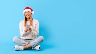 Noel Baba şapkalı gülümseyen genç kadın bağdaş kurmuş ve akıllı telefon kullanıyor. Hoş bir sürpriz gibi görünüyor. Mavi arka planda, boş alanda, afişte.