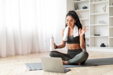 Neşeli genç Hintli kadın, evdeki online antremandan sonra su içiyor, atletik milenyum bayanı yoga minderinde oturuyor, şişeyi suyla tutuyor, dizüstü bilgisayara el sallıyor, teşekkürler fitness eğitmeni