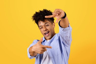 Gülümseyen kıvırcık saçlı siyah adam, parlak sarı arka planda odak ve yaratıcılığı sembolize eden elleriyle kamera çerçevesi yapıyor.