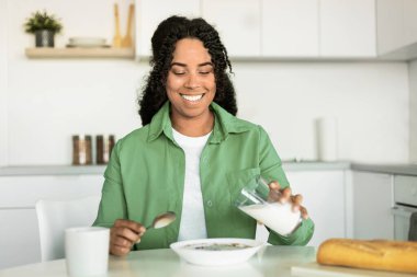 Lezzetli Kahvaltı. Neşeli genç siyah bayan bardaktan bardağa mısır gevreğiyle taze süt ekliyor, modern mutfakta sağlıklı bir yemek yiyor, sabahları, masada oturuyor. Beslenme kavramı