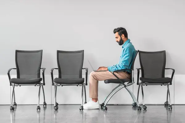Concentrated indian man using laptop while sitting in chair, preparing for job interview in quiet and empty waiting area, symbolizing focus and dedication, side view