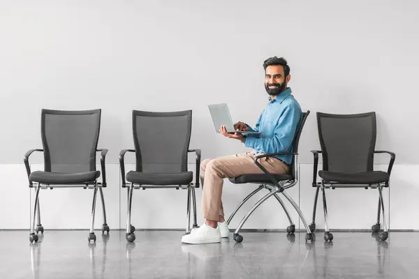 Happy indian male job candidate using laptop while sitting in waiting room, displaying relaxed and confident attitude before an interview, side view