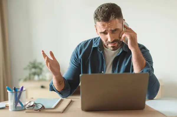 不满的中年男人拿着智能手机 一边坐在家里的办公桌旁一边用手机说话 一边在电脑上排解问题 多任务和商业职业 — 图库照片