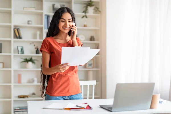 Energetic Millennial Eastern Woman Balancing Calls and Paper Work At Home Office. Engaged and in control, lady manages telephone conversation while working on laptop, work-life balance, copy space