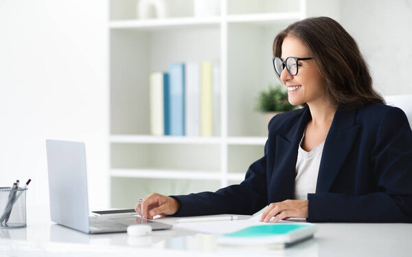Positive mature woman manager wearing formal outfit and eyeglasses sitting at desk, using laptop at modern office, sending emails, working on report, copy space