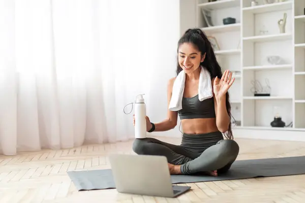 Joyful young indian woman drink water after online workout at home, athletic well-fir millennial lady sitting on yoga mat, hold bottle with water, waving at laptop screen, thanks fitness instructor
