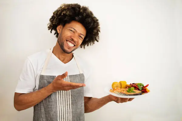 Smiling black man in apron holds a plate of salmon, showcasing healthy meal prep in modern kitchen setting. Chef presenting tasty fish dish, embodying lifestyle of wellness and weight loss