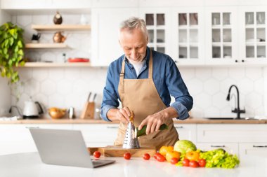 Önlüklü kıdemli adam mutfakta yemek pişirirken video dersi izliyor, bilgisayar kullanan ve evde sağlıklı öğle yemeği ya da akşam yemeği hazırlayan, sağlıklı beslenmenin tadını çıkaran yaşlı adam.