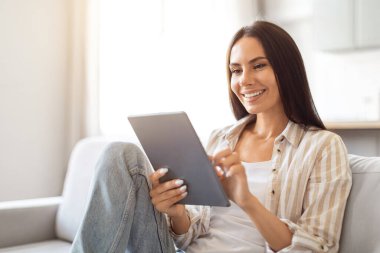 Harika bir uygulama. Dijital tabletli mutlu genç bayan, evinde oturuyor, gülümseyen milenyum kadını oturma odasında modern Gadget ile dinleniyor, internete bakıyor ya da internetten alışveriş yapıyor,