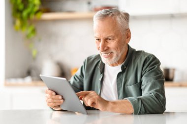 Mutfaktaki masada dinlenirken dijital tablet kullanan, evde modern Gadget 'ta gülümseyen, internetten alışveriş yapan, emekliliğin tadını çıkaran yaşlı adam.
