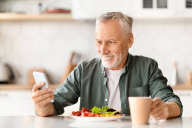 Mutfakta kahvaltı yaparken akıllı telefon kullanan mutlu kıdemli adam, cep telefonuyla interneti karıştıran, haberleri okuyan ya da yemek yerken sosyal medyada fotoğrafları kontrol eden yaşlı beyefendi.