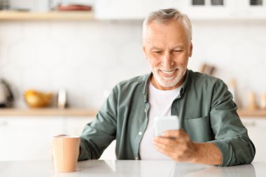 Gülümseyen, akıllı telefon kullanan ve mutfakta kahve içen yaşlı bir adamın portresi, mutlu, yakışıklı, yaşlı ve cep telefonuyla evde dinlenen, online haberler okuyan ve sıcak içeceğin tadını çıkaran bir beyefendi.