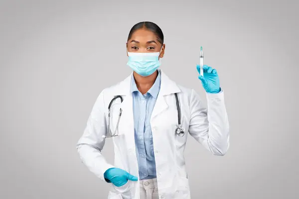 Smiling millennial black lady doctor in white coat, protective gloves, mask show syringe, isolated on gray background studio. Vaccination recommendation, medicine, health care in clinic