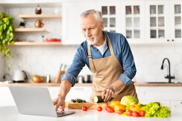 Smiling Elderly Man Looking Recipe On Laptop While Cooking Food In Kitchen, Happy Senior Gentleman Using Computer, Attending Culinary Class And Preparing Healthy Vegetarian Lunch At Home, Copy Space