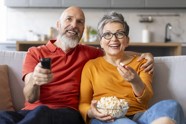 Portrait Of Senior Spouses Watching Tv At Home And Eating Popcorn, Happy Elderly Husband And Wife Relaxing On Couch In Living Room And Embracing, Man Holding Remote Controller, Closeup