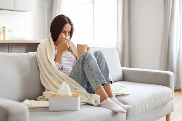 Cold And Flu Concept. Sick young woman blowing her running nose in paper tissue and drinking tea at home, ill female covered in blanket sitting on couch in living room, feeling unwell, copy space