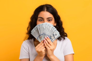Mutlu Hintli bir kadının portresi. Elinde bir sürü para banknotları ve kameraya bakıyor. Sarı arka planda izole edilmiş nakit dolarların arkasına saklanıyor.