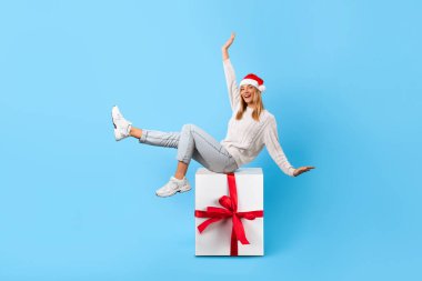 Noel Baba şapkalı neşeli genç bir kadın kırmızı kurdeleli büyük beyaz hediye kutusunda mutlu bir şekilde poz veriyor.