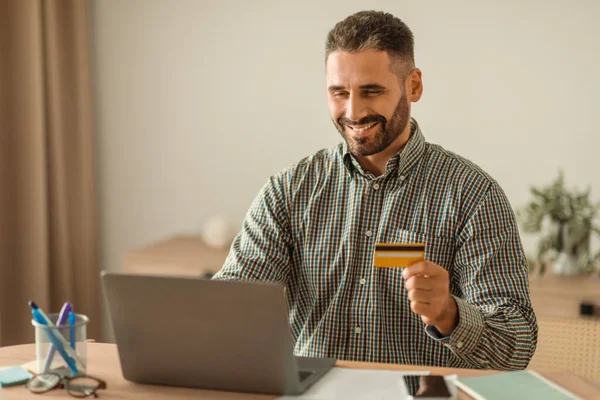 European man holding credit card while shopping online on laptop computer in home office indoor, exploring internet stores and bank offers, making transactions with happy smile