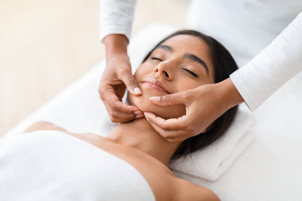 Beautiful Young Indian Woman Getting Face Lifting Massage In Luxury Spa Salon, Masseur Making Double Chin Treatment Procedure To Relaxed Lady Lying With Eyes Closed, Closeup Portrait