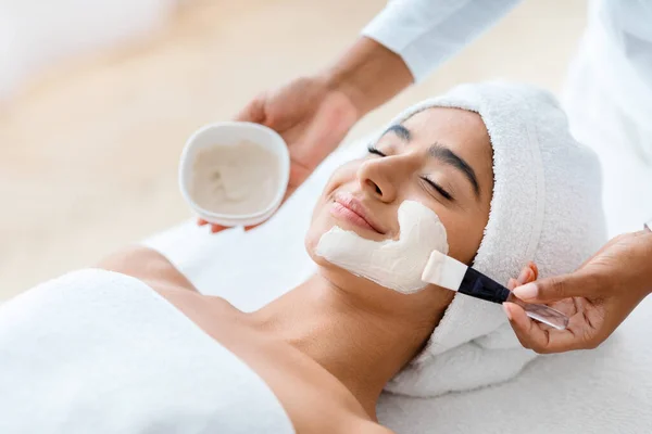 Anti-Aging Skincare. Cosmetologist applying clay mask on face of young indian woman, beautiful eastern lady with towel on head lying on table, enjoying beauty treatments in spa salon, closeup