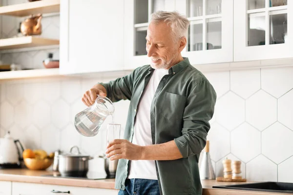 Portrait of happy elderly man filling his glass with water from jug in kitchen at home, smiling senor gentleman pouring healthy drink, taking care about body hydration and wellness, copy space