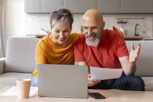 Portrait of excited senior spouses with papers and laptop celebrating good news at home, happy elderly couple reading mail and enjoying success, reacting to credit approvance or financial profit