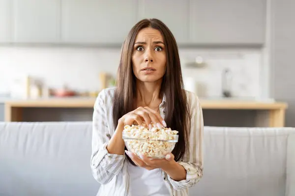Frightened young woman watching scary movie at home and eating popcorn, scared millennial female sitting on couch in living room and looking at tv screen, emotionally reacting to shocked content