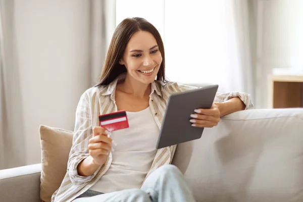 Online Shopping Concept. Smiling Beautiful Female Using Digital Tablet And Credit Card While Relaxing On Couch In Cozy Living Room At Home, Happy Young Lady Purchasing In Internet, Copy Space