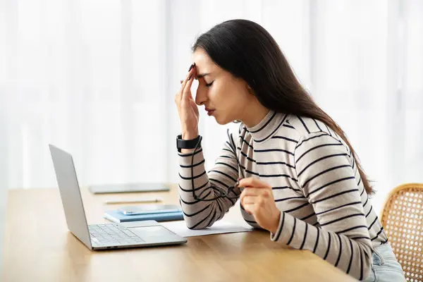 Sad young arab woman work at computer, suffer from migraine, health problems, pain and headache, feeling bad in living room interior. Stress, mistake, deadline at laptop at home