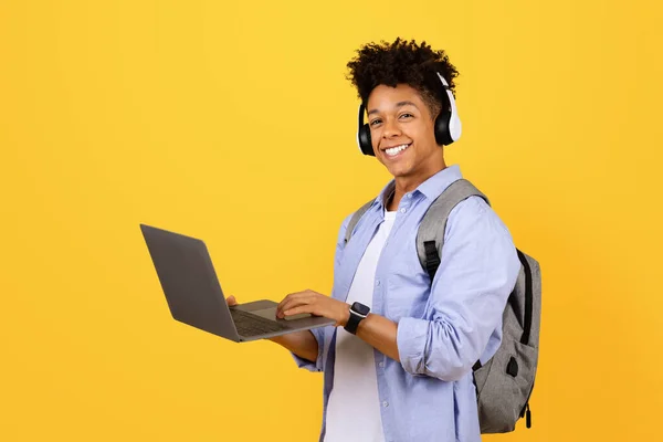 Smiling black male student wearing headphones and backpack holds laptop computer, ready for online learning on bright yellow background