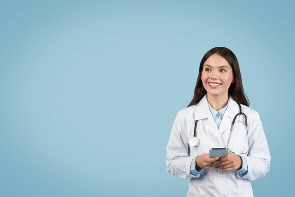 Happy female doctor in white lab coat holding smartphone looks upward at free space, poised with stethoscope around her neck against soft blue backdrop
