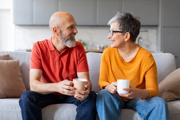 Happy Senior Couple Drinking Coffee And Relaxing On Couch In Living Room, Cheerful Elderly Spouses Resting Tv At Home And Enjoying Hot Drinks, Spending Weekend Time Together, Closeup