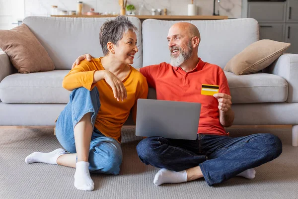 Online Banking. Happy Older Couple Using Laptop And Credit Card At Home, Cheerful Senior Spouses Making Payments In Internet While Sitting On Floor In Living Room, Smiling To Each Other