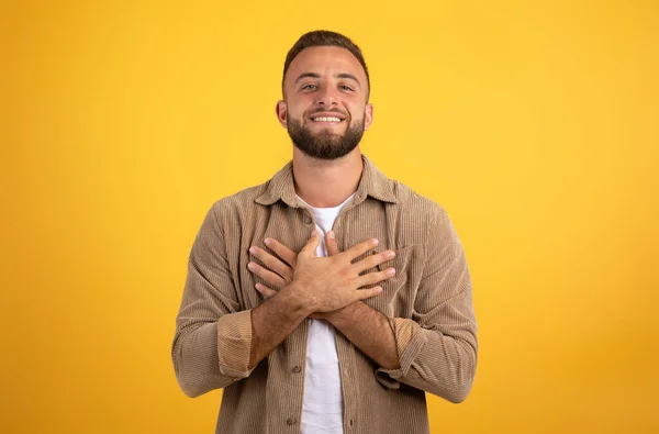 Glad handsome young european man presses hands to chest, isolated on yellow studio background. Thanks gesture, gratitude sign, love, people emotions and lifestyle, good news