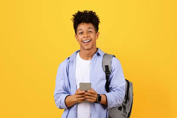快乐的年轻黑人男生 背着背包 舒舒服服地使用智能手机 站在明亮的黄色背景下对着摄像机微笑 — 图库照片