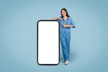 Tıp üniformalı genç bayan büyük telefona yaslanıyor, tıbbi reklamlar için ekranı işaret ediyor, mavi arka planda sağlık iletişimi, modelleme