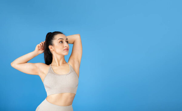 A good-looking lady with brown hair, in comfy beige sports gear, stands against a blue background, her in-shape body is a sign of her active life and love for sports, looking at copy space