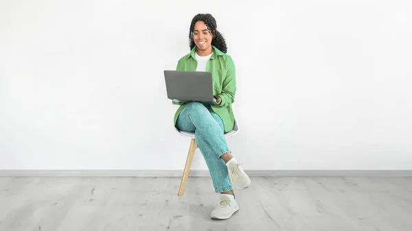 Smiling black woman sits with laptop computer against white wall, wearing casual green shirt, immersed in remote job, showcasing technology and career. Modern freelance work. Panorama, copy space